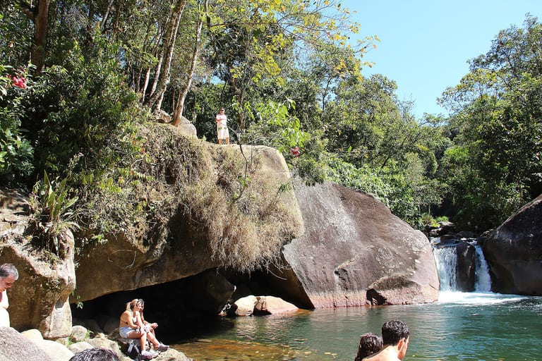 Pedra alta usada para saltos ao lado do Poção da Maromba, com banhistas no poço de água verde, em Visconde de Mauá