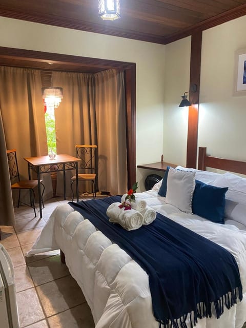 Suíte da Pousada do Figa com cama de casal arrumada, toalhas dobradas e mesa para dois junto à janela