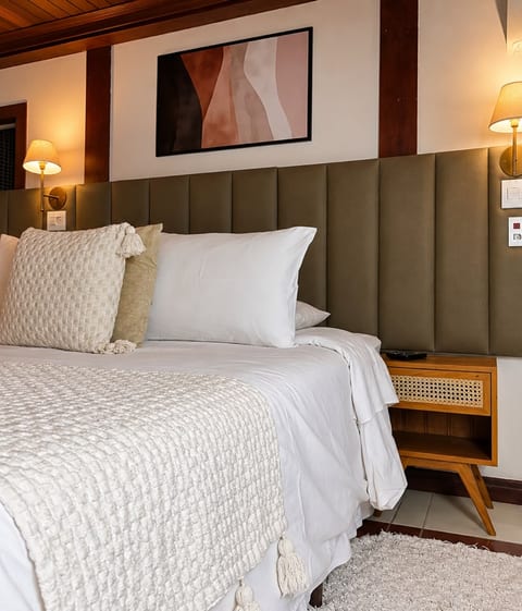 Cama queen com manta de tricô, almofadas com pompom e criado-mudo de palhinha na Suíte 1 da Pousada do Figa