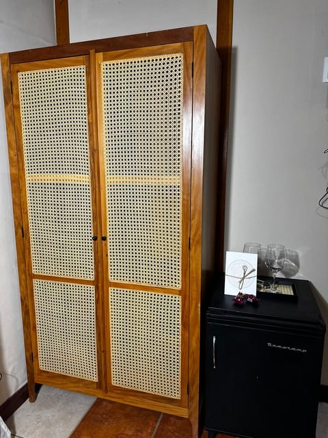 Guarda-roupa de madeira e palhinha, frigobar e taças com bilhete de boas-vindas na Suíte 2 da Pousada do Figa